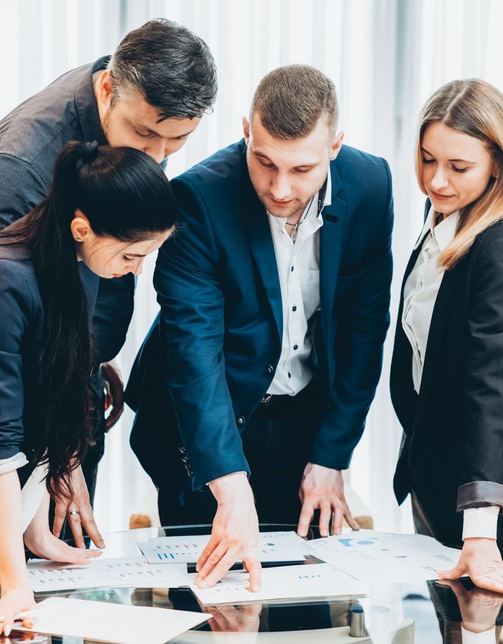 Quattro professionisti riuniti attorno a un tavolo con documenti e grafici stampati, uno indica un punto preciso: il coordinamento che porta a termine ciò che è stato deciso.